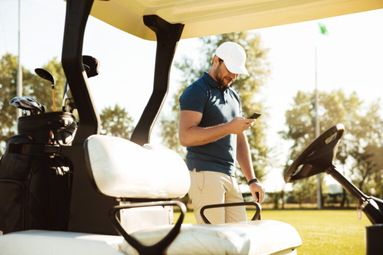Young man using mobile phone while standing at a golf cart on a field